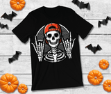 Black t-shirt with a skeleton wearing a red cap on a wooden surface with Halloween decorations.