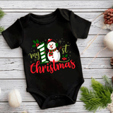 Black baby onesie with a Christmas design featuring a snowman and text on a wooden surface.