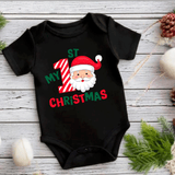 Black baby onesie with Santa Claus design and text on a wooden surface with Christmas decorations.