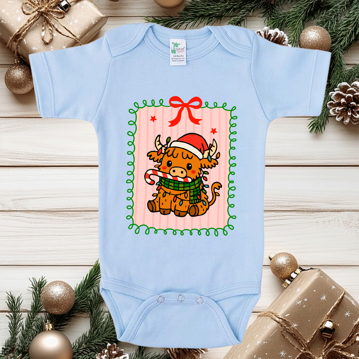 Light blue baby onesie with a Christmas cow design on a wooden surface with holiday decorations.