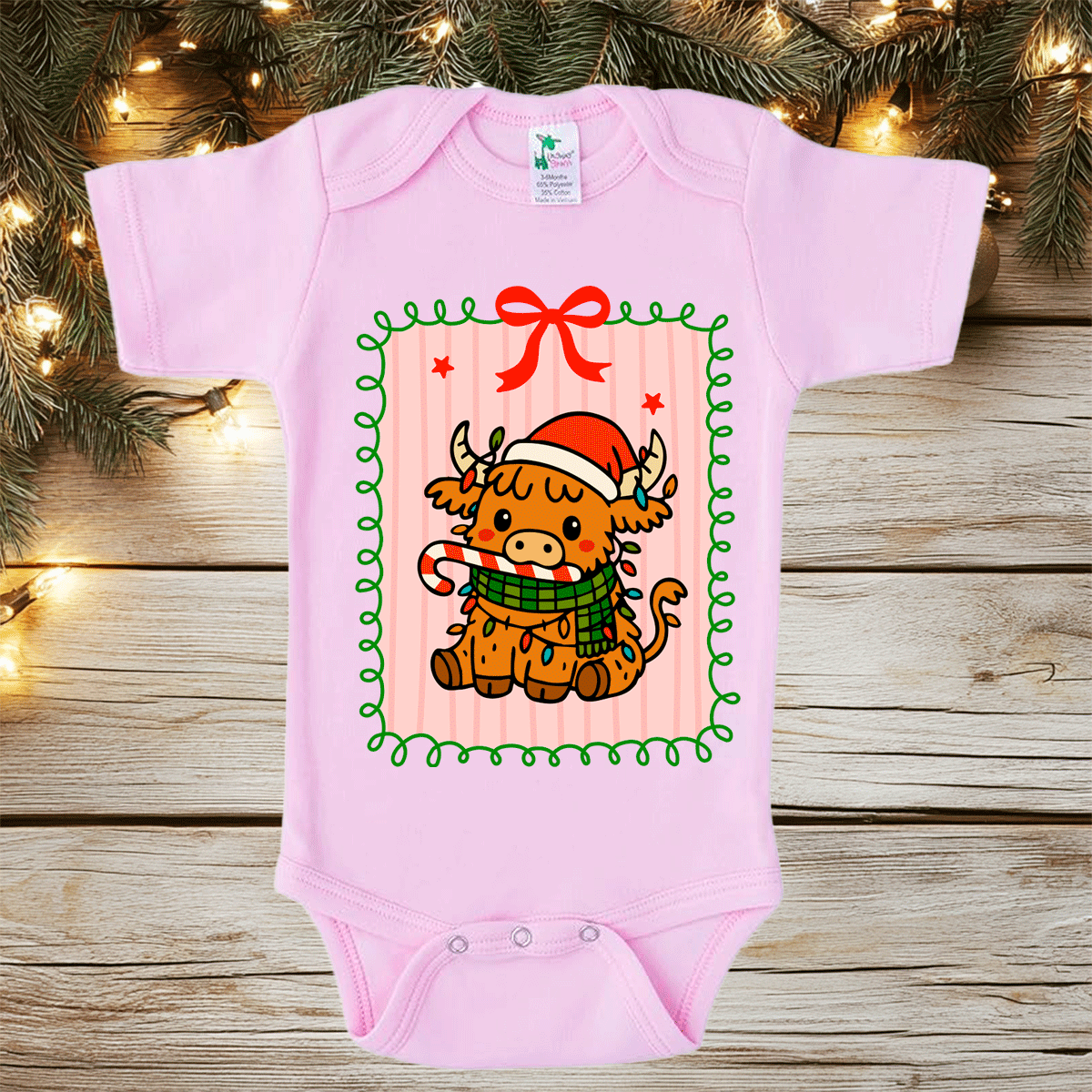Pink baby onesie with a Christmas-themed design of a cow wearing a Santa hat and scarf, placed on a wooden surface with festive lights and decorations.