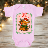 Pink baby onesie with a Christmas-themed design of a cow wearing a Santa hat and scarf, placed on a wooden surface with festive lights and decorations.