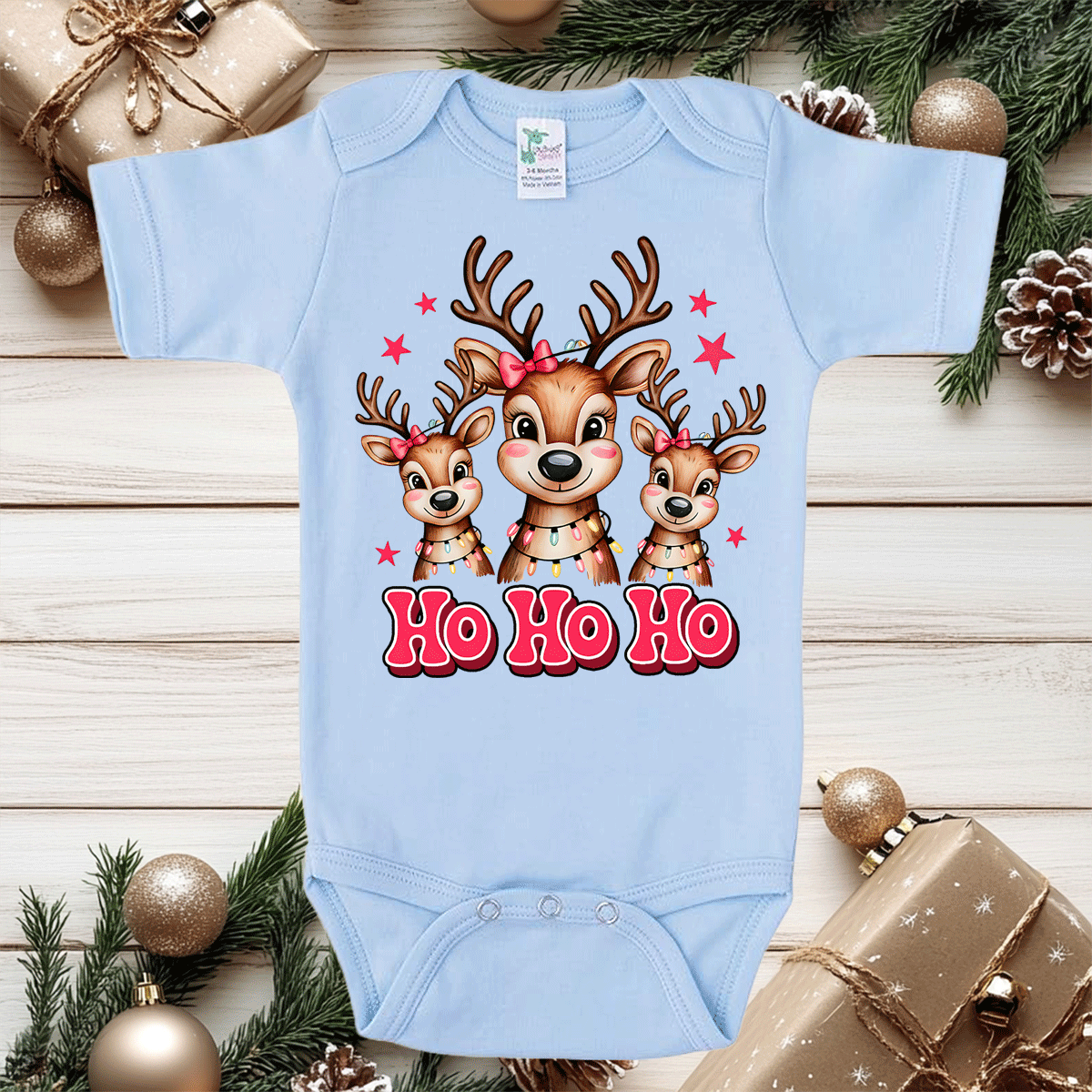 Light blue baby onesie with reindeer design and 'Ho Ho Ho' text on a wooden surface with Christmas decorations.