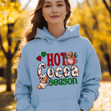 Person wearing a light blue hoodie with 'Hot Cocoa Season' text and graphics in an autumn setting.