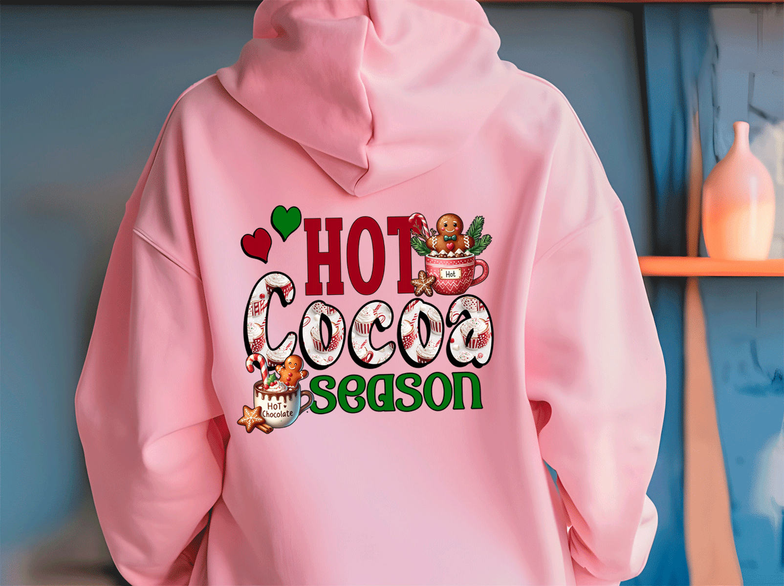 Pink hoodie with 'Hot Cocoa Season' text and graphics on a blue wall background