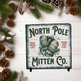 Decorative sign for North Pole Mitten Co. with green mittens and pinecones on a wooden background