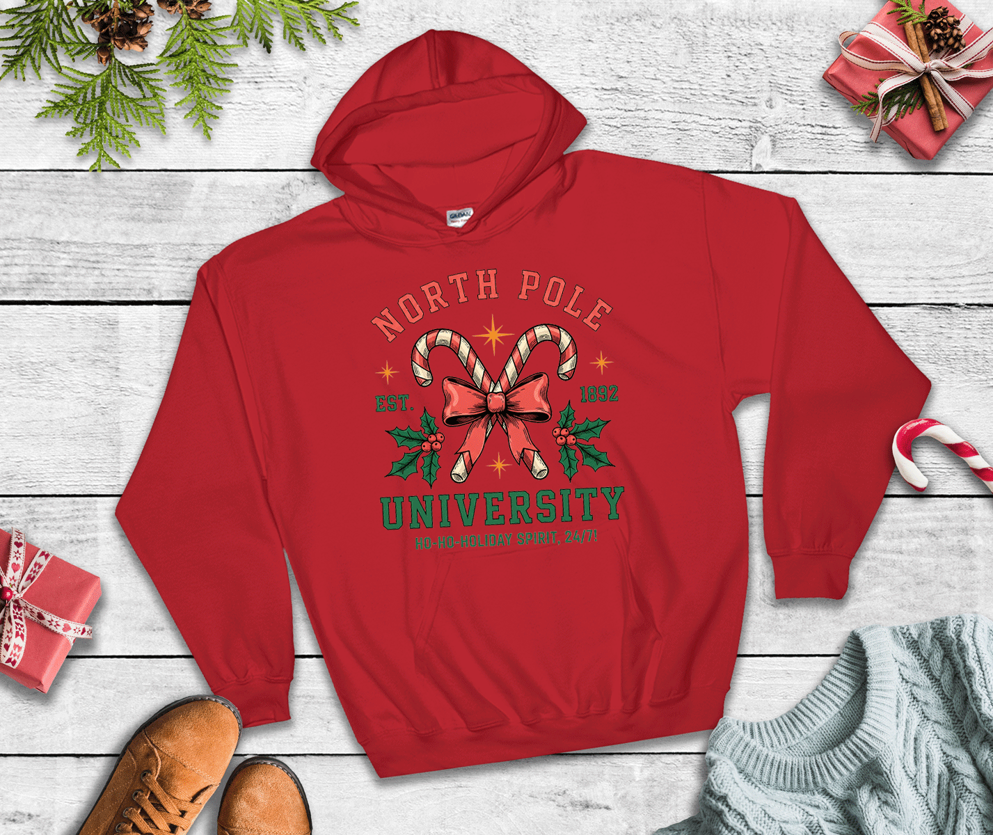 Red hoodie with 'North Pole University' design on a wooden surface with Christmas decorations.