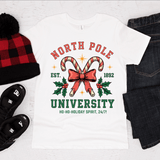 White t-shirt with 'North Pole University' design, surrounded by festive items on a light background