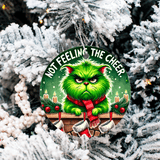 Christmas ornament with a green cat wearing a red scarf, sitting on a sleigh with candles and text 'Not Feeling the Cheer' against a snowy tree background.