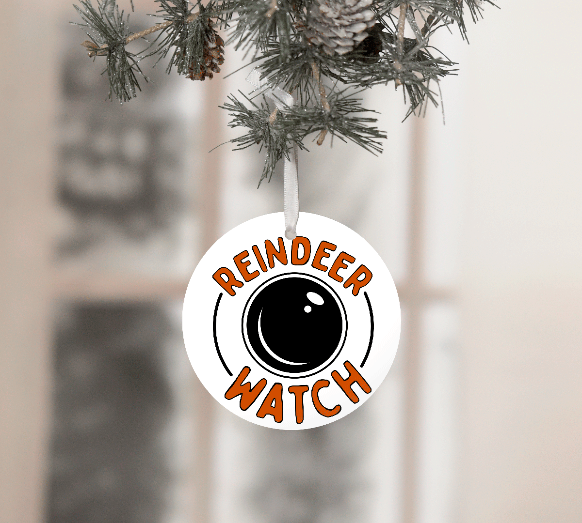 Round ornament with 'Reindeer Watch' text hanging on a Christmas tree.