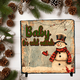 Decorative sign with a snowman and 'Baby it's cold outside' text, surrounded by pinecones and branches.