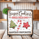 Decorative sign with Christmas-themed text and cookies on a wooden surface.